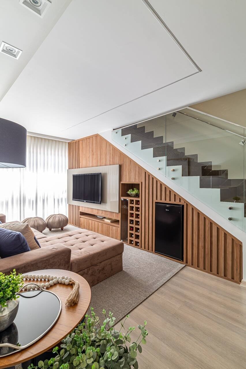 Phòng khách hiện đại với ghế sofa và TV, cầu thang kính, nội thất gỗ tinh tế và cây xanh.