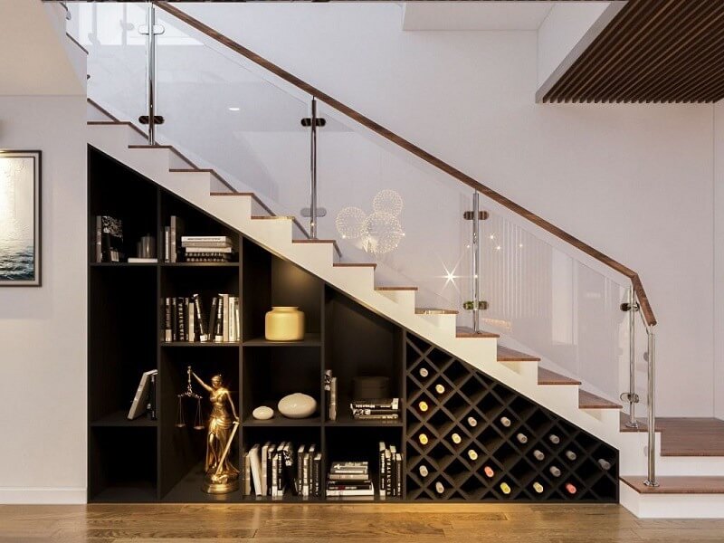 Giá sách dưới cầu thang với kệ trưng bày sách, chai rượu, và đồ trang trí trong không gian hiện đại và sáng sủa.