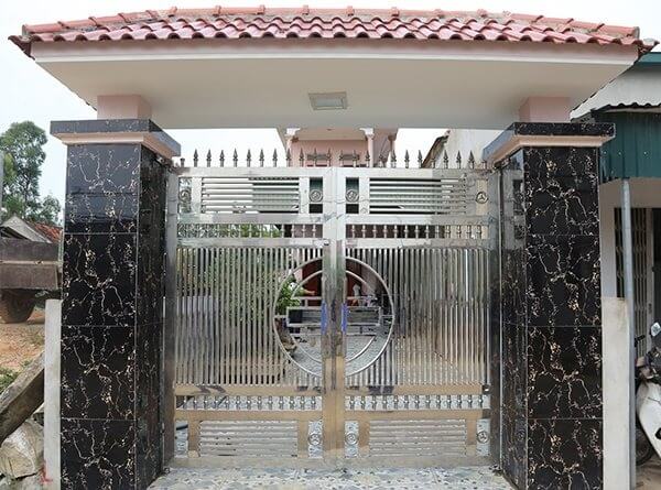 Cổng nhà bằng thép không gỉ với mái ngói đỏ và cột ốp đá tối màu, thiết kế hiện đại và sang trọng.