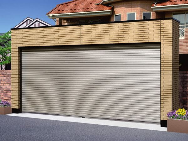 Cửa cuốn gara hiện đại, màu xám bạc, mặt tiền nhà gạch, mái ngói đỏ, cây cỏ trang trí hai bên.