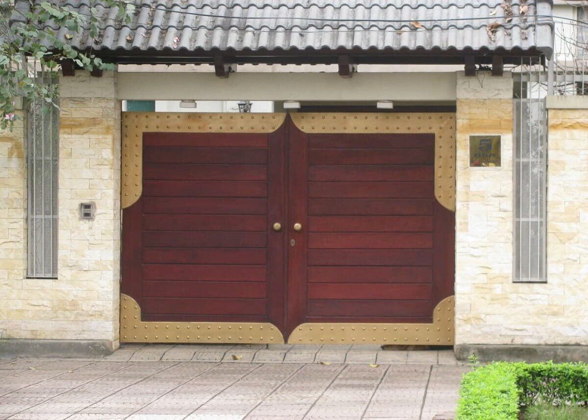 Cổng gỗ nâu đỏ lớn với viền kim loại vàng, mái ngói xám, xung quanh tường lát gạch sáng màu.