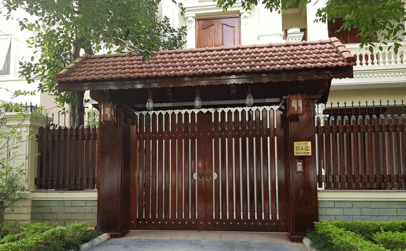 Cổng gỗ nâu sang trọng của biệt thự, mái ngói đỏ, kiến trúc truyền thống, bao quanh bởi cây xanh và hàng rào.