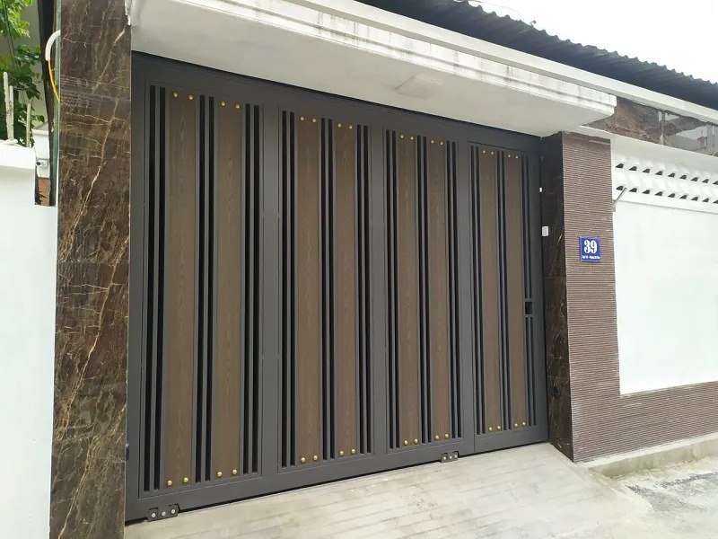 Cổng trượt kim loại màu nâu sẫm hiện đại, lắp đặt trước ngôi nhà với bảng số đường phố bên phải.