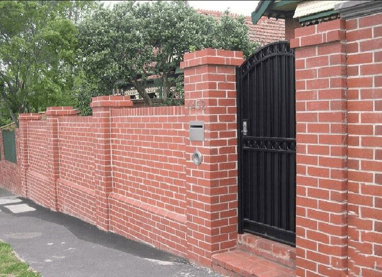 mau-cong-nha-xay-bang-gach-dep Tường gạch đỏ và cổng sắt đen trước ngôi nhà, có cây xanh phía sau.