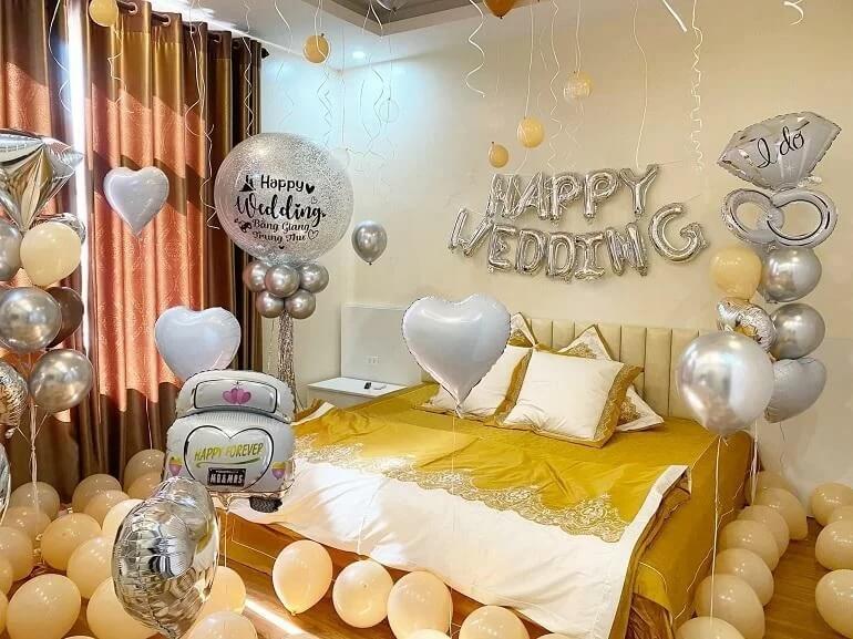 Trang trí phòng cưới với bóng bay trắng và vàng, chữ Happy Wedding trên tường, tạo không gian lãng mạn.