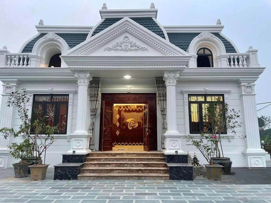 Biệt thự kiến trúc tân cổ điển trắng, cổng gỗ đỏ, cửa sổ kính, hai cây cảnh trước sân.