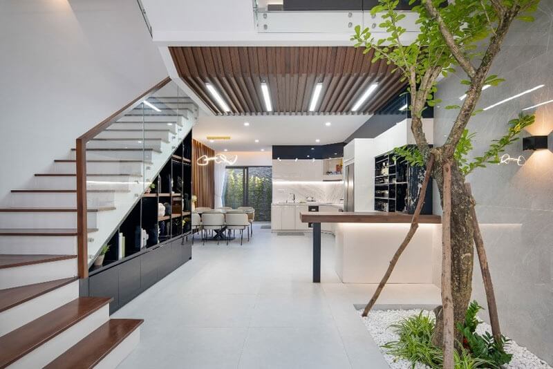 Phòng khách hiện đại với cầu thang gỗ, cây xanh và bếp mở rộng rãi, ánh sáng tự nhiên tràn ngập.