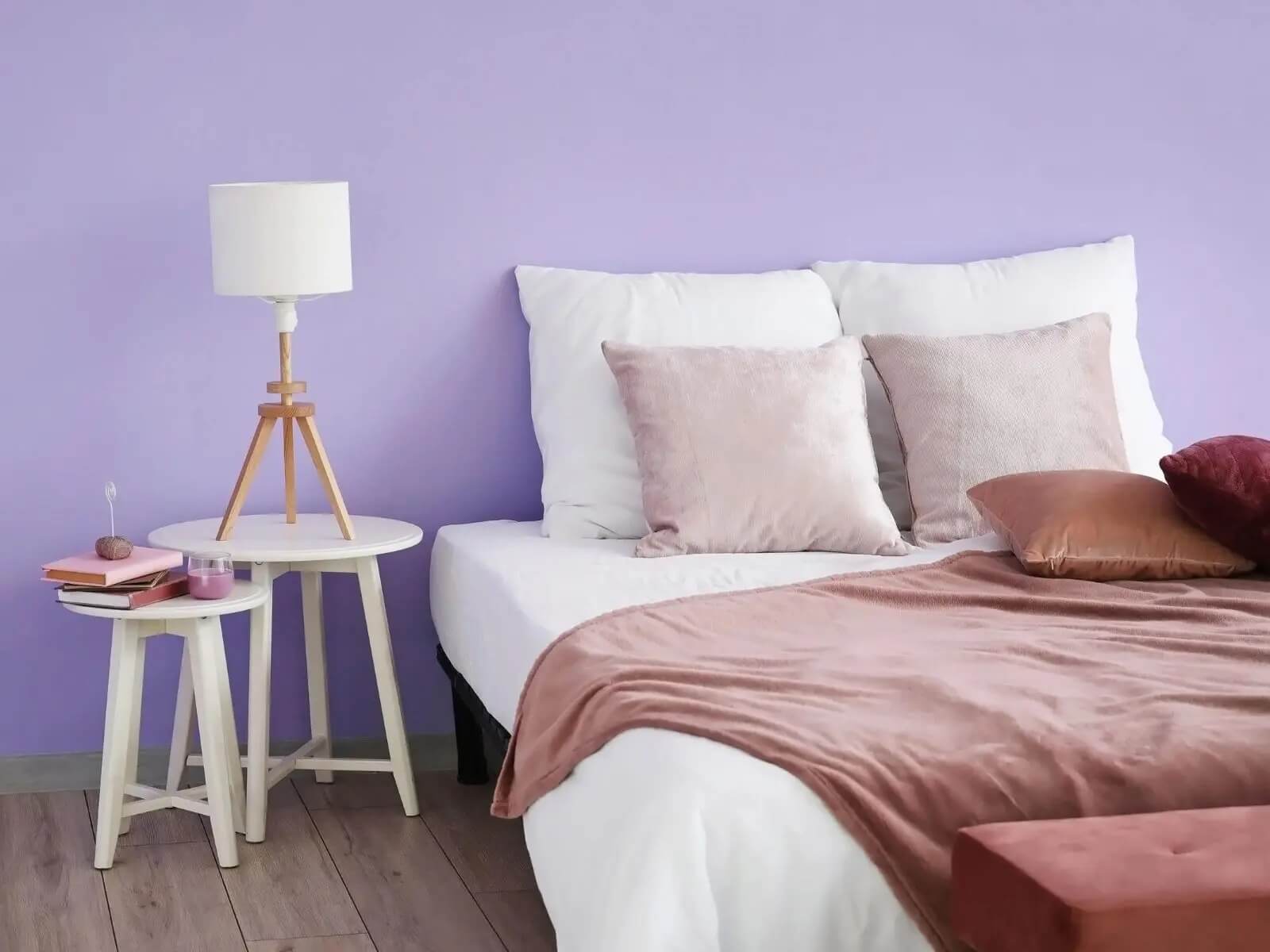Phòng ngủ với giường trắng, gối và ga màu hồng, tường màu tím, bàn nhỏ và đèn trang trí thanh lịch.