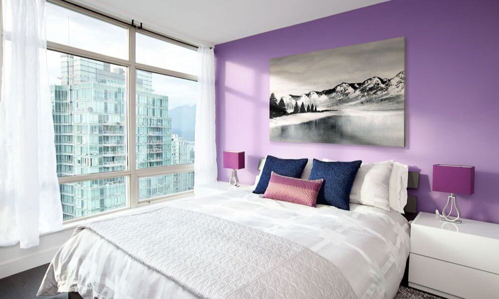 Phòng ngủ hiện đại với tường màu tím, giường đôi, cửa sổ lớn nhìn ra thành phố, và tranh phong cảnh treo tường.