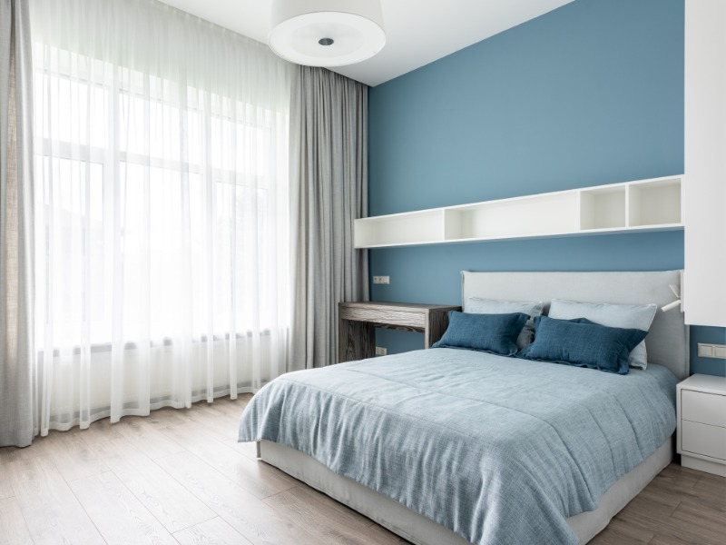 Phòng ngủ hiện đại với tường màu xanh, giường lớn và ánh sáng tự nhiên qua rèm cửa trắng.