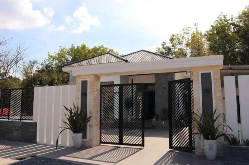 Nhà hiện đại với mái ngói và cổng sắt, bao quanh bởi cây xanh dưới bầu trời nhiều mây.