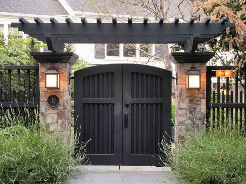 mau-thiet-ke-cong-nha-vuon-dep-3 Cổng gỗ màu đen với mái vòm treo, trang trí đèn và cây xanh xung quanh, trước một ngôi nhà.