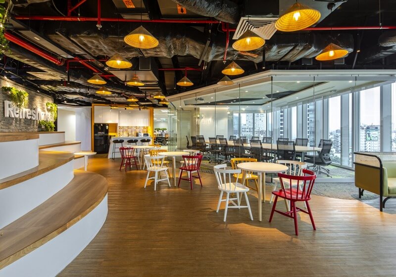 Không gian văn phòng hiện đại với bàn ghế màu sắc bên dưới ánh đèn vàng, cửa sổ kính lớn nhìn ra thành phố.