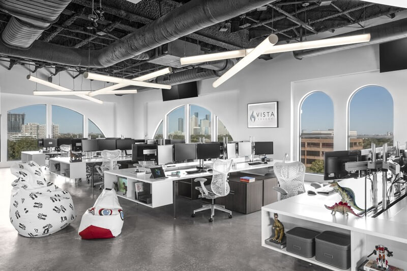 Văn phòng hiện đại với chỗ ngồi mở, nhiều máy tính và ghế công thái học, cửa sổ lớn nhìn ra thành phố.