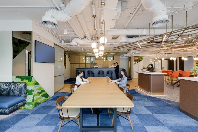 Văn phòng hiện đại với nội thất sáng sủa, bàn làm việc gỗ dài và nhân viên đang sử dụng laptop.