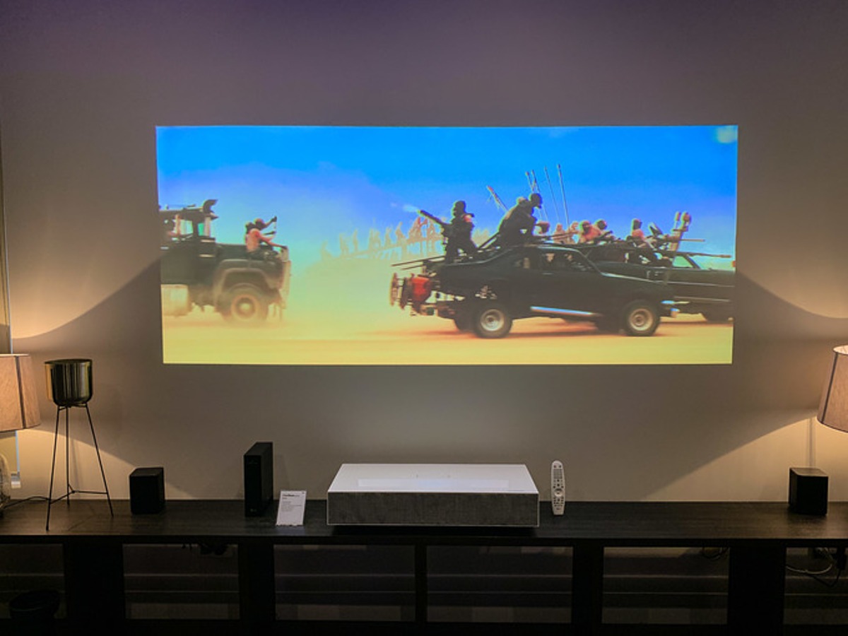 Máy chiếu hiển thị cảnh hành động phim trên tường, trong phòng khách với đèn và bàn.