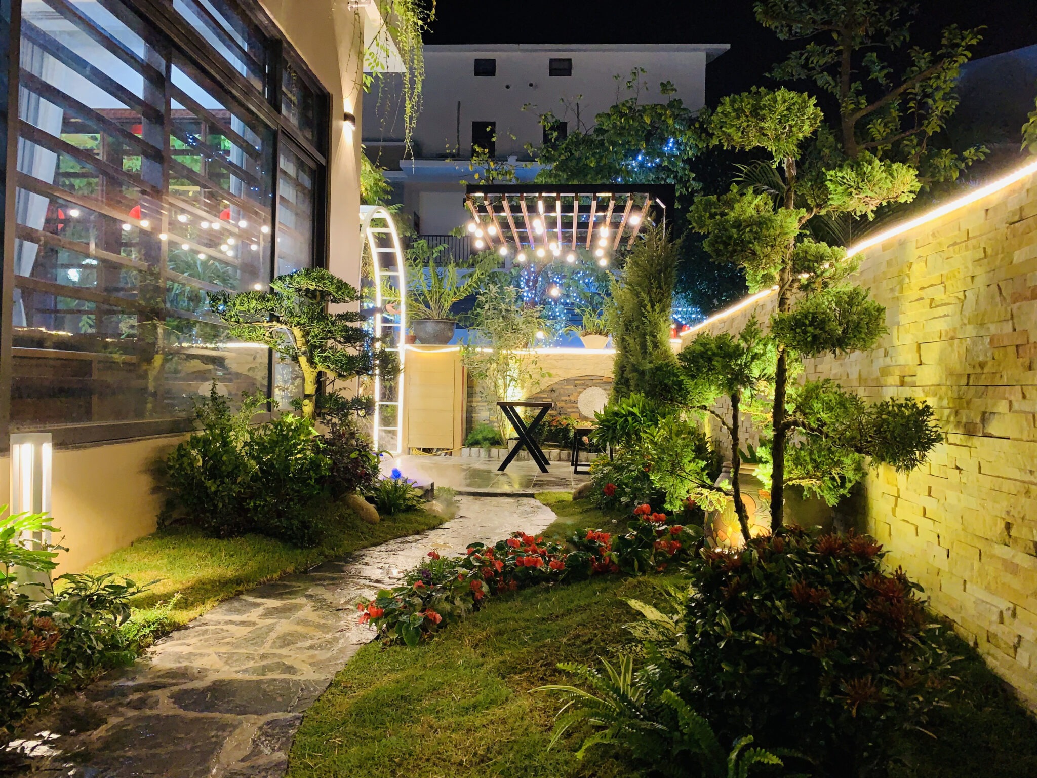 Lối đi sân vườn với đèn sáng và cây xanh, bàn ghế thư giãn, tạo không gian ấm áp về đêm.