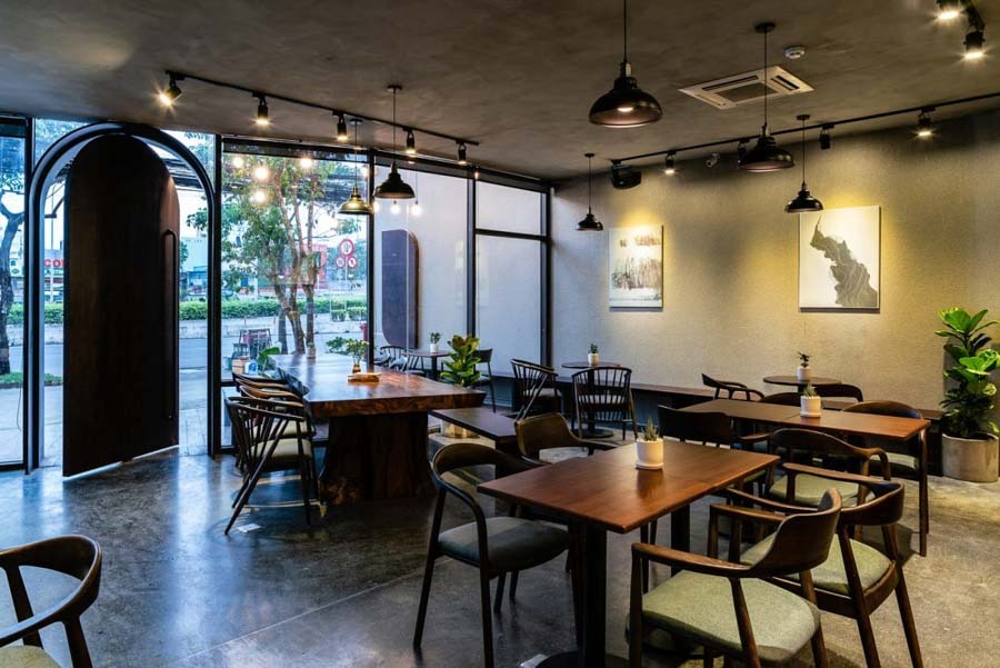 Quán cà phê hiện đại với nội thất gỗ, cửa kính lớn và cây xanh, tạo không gian thoáng mát và sáng sủa.