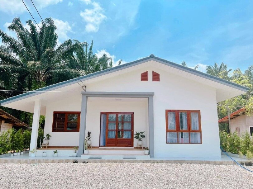 nha-2-mai-truoc-sau-don-gian-4 Ngôi nhà cấp 4 mái lệch màu trắng với cửa gỗ, cây xanh và bầu trời trong xanh.