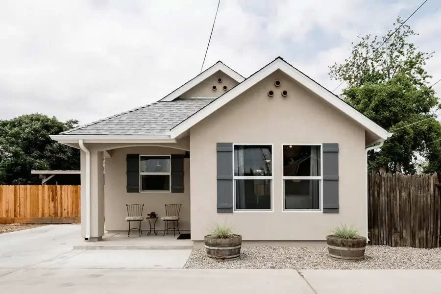 Nhà nhỏ kiểu cottage với mái xanh, hàng rào gỗ và sân rộng. Cảnh yên bình và cây xanh phía sau.