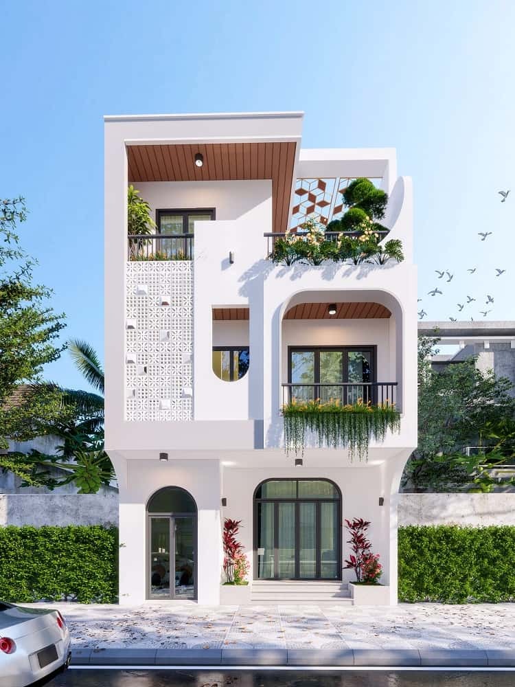 Nhà phố ba tầng hiện đại với ban công cây xanh và tường trắng, phía trước có ô tô đậu trên phố.