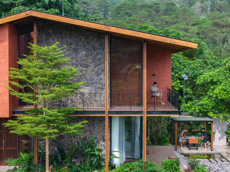 Ngôi nhà hiện đại giữa thiên nhiên xanh, có ban công và người ngồi đọc sách, phía dưới là bàn ghế ngoài trời.