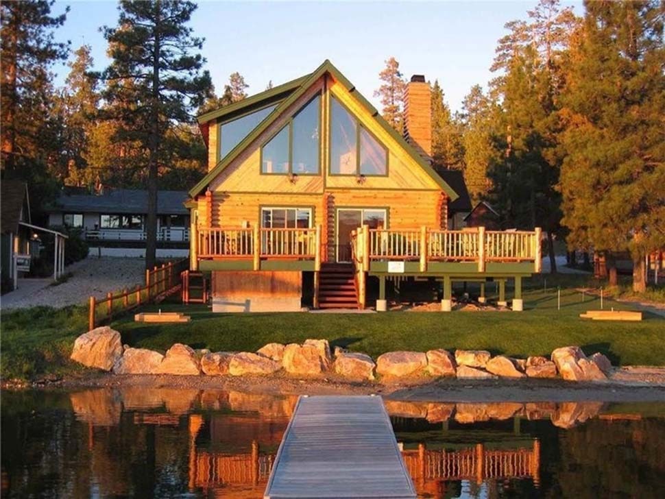 Ngôi nhà gỗ bên hồ với ban công và cửa sổ lớn, được bao quanh bởi cây cối và mặt trời lặn.