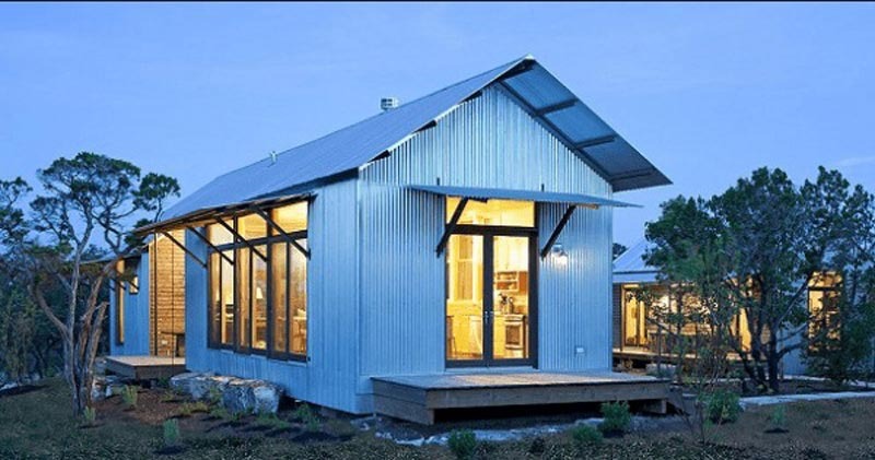 Nhà nhỏ bằng kim loại với cửa sổ rộng, mái dốc, tọa lạc giữa thiên nhiên xanh tươi vào buổi hoàng hôn.