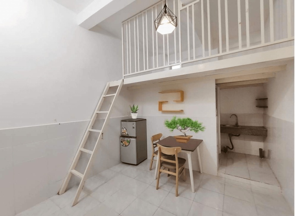 Căn phòng nhỏ gác lửng với bàn ăn, ghế, thang gỗ, tủ lạnh và đèn treo hiện đại.