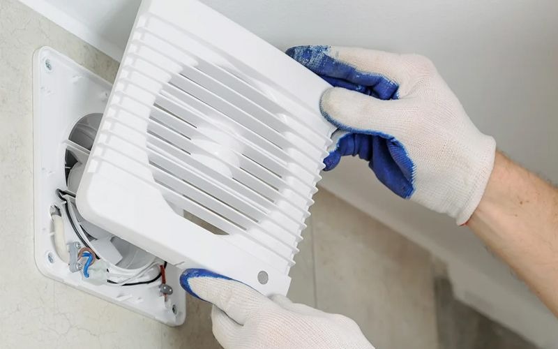 Nhân viên đang lắp đặt quạt thông gió trên tường với găng tay trắng, cải thiện thông khí cho không gian.