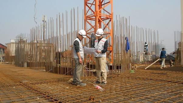 Công nhân công trường xây dựng đội mũ bảo hộ kiểm tra bản vẽ tại công trình với cần trục và cột thép xung quanh.
