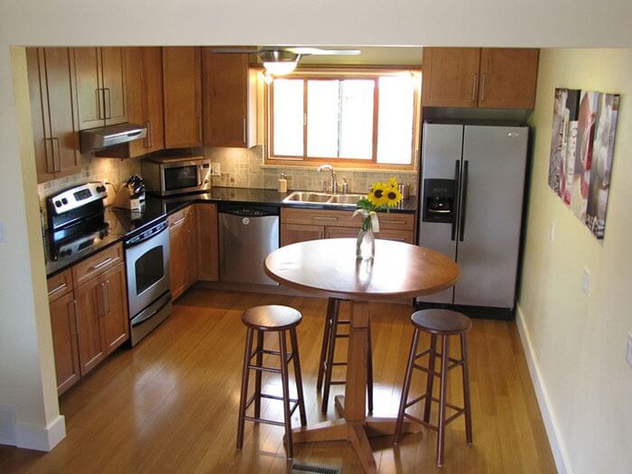 Nhà bếp hiện đại với tủ gỗ, bàn tròn có hoa hướng dương và ghế quầy, sáng tự nhiên từ cửa sổ.