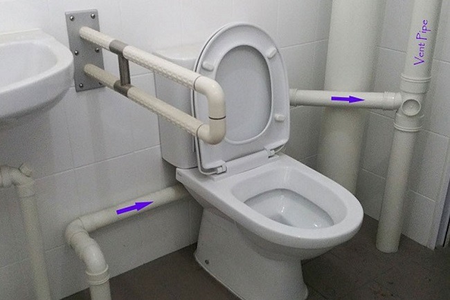 Nhà vệ sinh với ống dẫn nước phức tạp và tay vịn hỗ trợ, thiết kế không hợp lý.