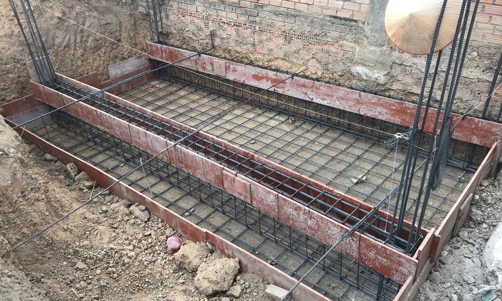 phan-loai-mong-bang-2-phuong-2 Nền móng công trình xây dựng với thép cốt bê tông, gạch lát và nón lá treo bên tường.