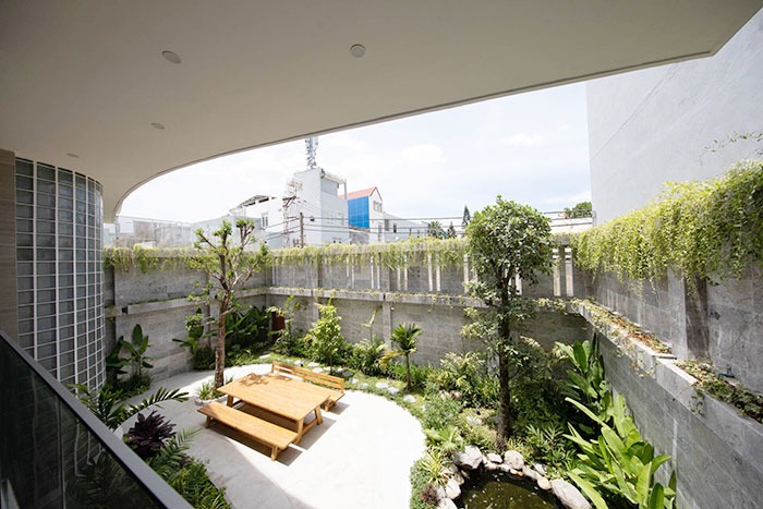 Khu vườn xanh mát với bàn gỗ ngoài trời, cây cối và ao nhỏ, tạo không gian thư giãn giữa tường xi măng.