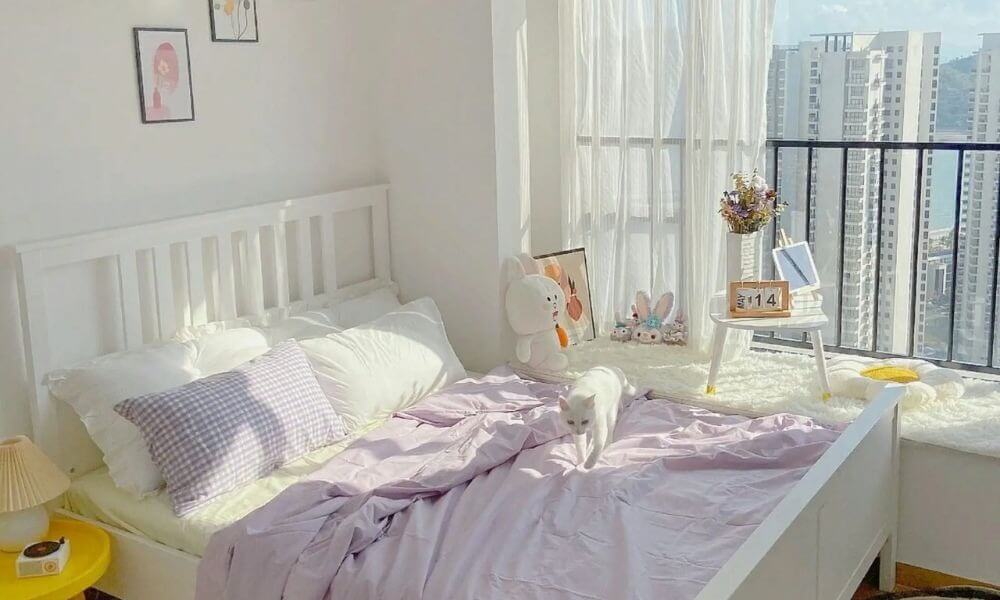 Phòng ngủ sáng sủa với giường trắng, ga trải màu tím nhạt, mèo trắng; cửa sổ lớn, tầm nhìn thành phố.