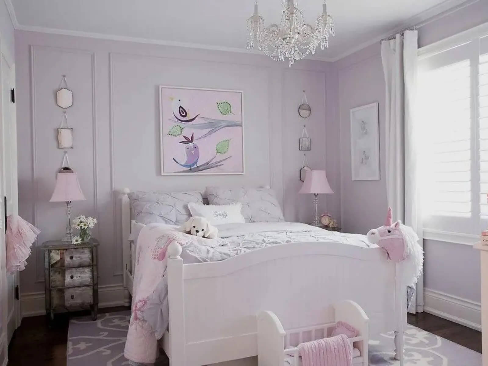 Phòng ngủ trẻ em màu tím pastel với đèn chùm pha lê, giường trắng và đồ chơi kỳ lân hồng bên cạnh.