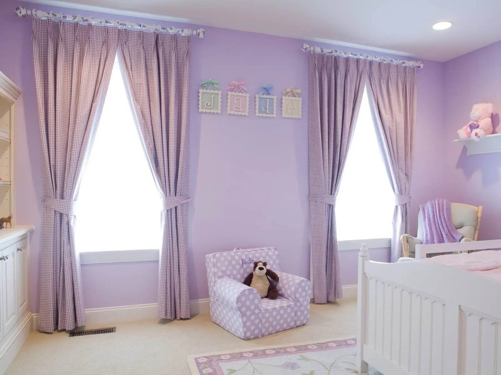 Phòng ngủ trẻ em màu tím với rèm cửa kẻ ô, giường cũi trắng và ghế bành họa tiết chấm bi.