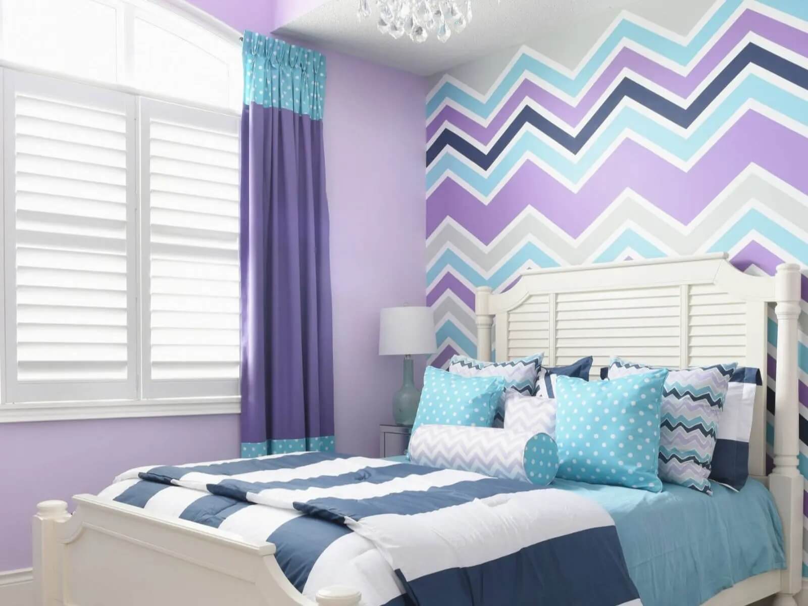 Phòng ngủ hiện đại với tường sọc zigzag tím và xanh, giường trắng, gối hoạ tiết chấm bi trên nền sáng.