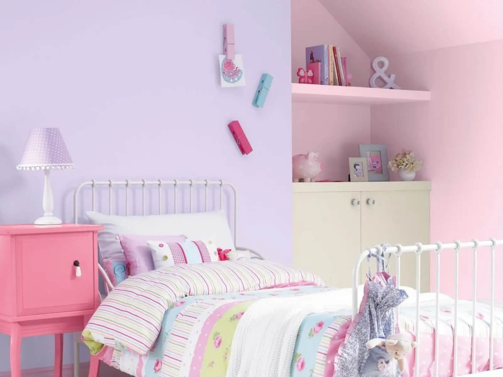 Phòng ngủ trẻ em tông hồng, giường sắt trắng, tủ đầu giường màu hồng và kệ sách trang trí.