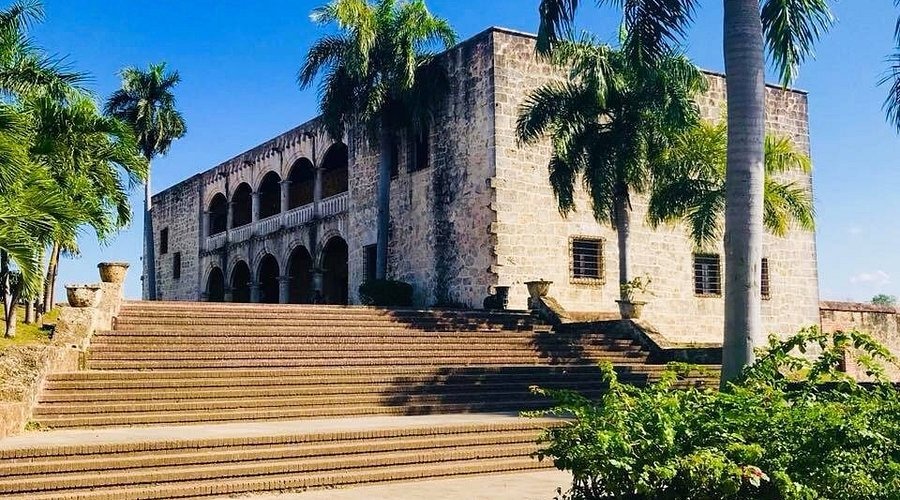 Tòa nhà lịch sử với cầu thang đá và cây cọ, bầu trời xanh nền trời sáng.