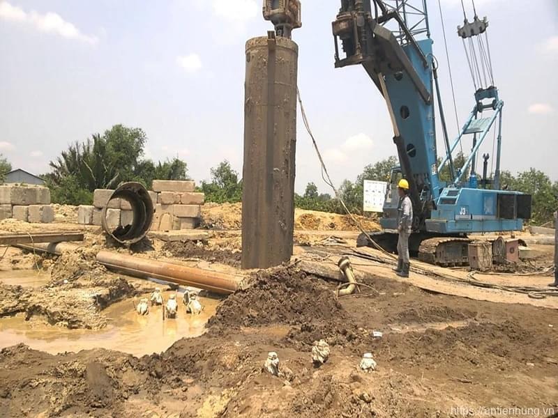 Công nhân đang giám sát máy khoan tại công trường xây dựng với nền đất sét và máy móc hạng nặng.