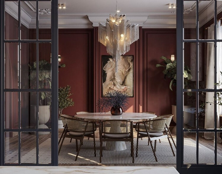 Phòng ăn sang trọng với bàn tròn, đèn chùm lộng lẫy và tường màu đỏ đậm, cây xanh trang trí xung quanh.
