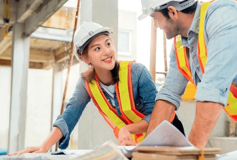 Kỹ sư xây dựng thảo luận bản vẽ tại công trường, mặc áo phản quang và đội mũ bảo hộ.