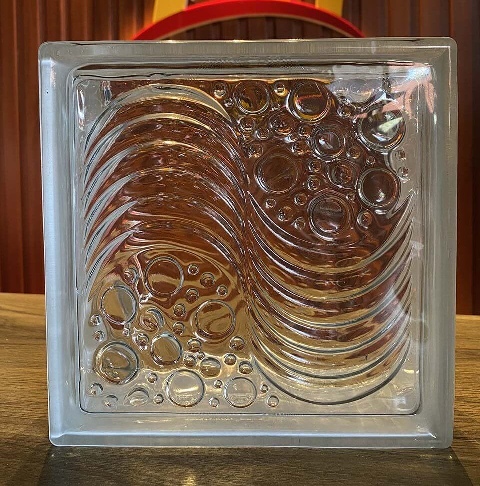 Khối kính nghệ thuật với hoa văn sóng và bọt, đặt trên bàn gỗ, ánh sáng chiếu qua tạo hiệu ứng đẹp mắt.