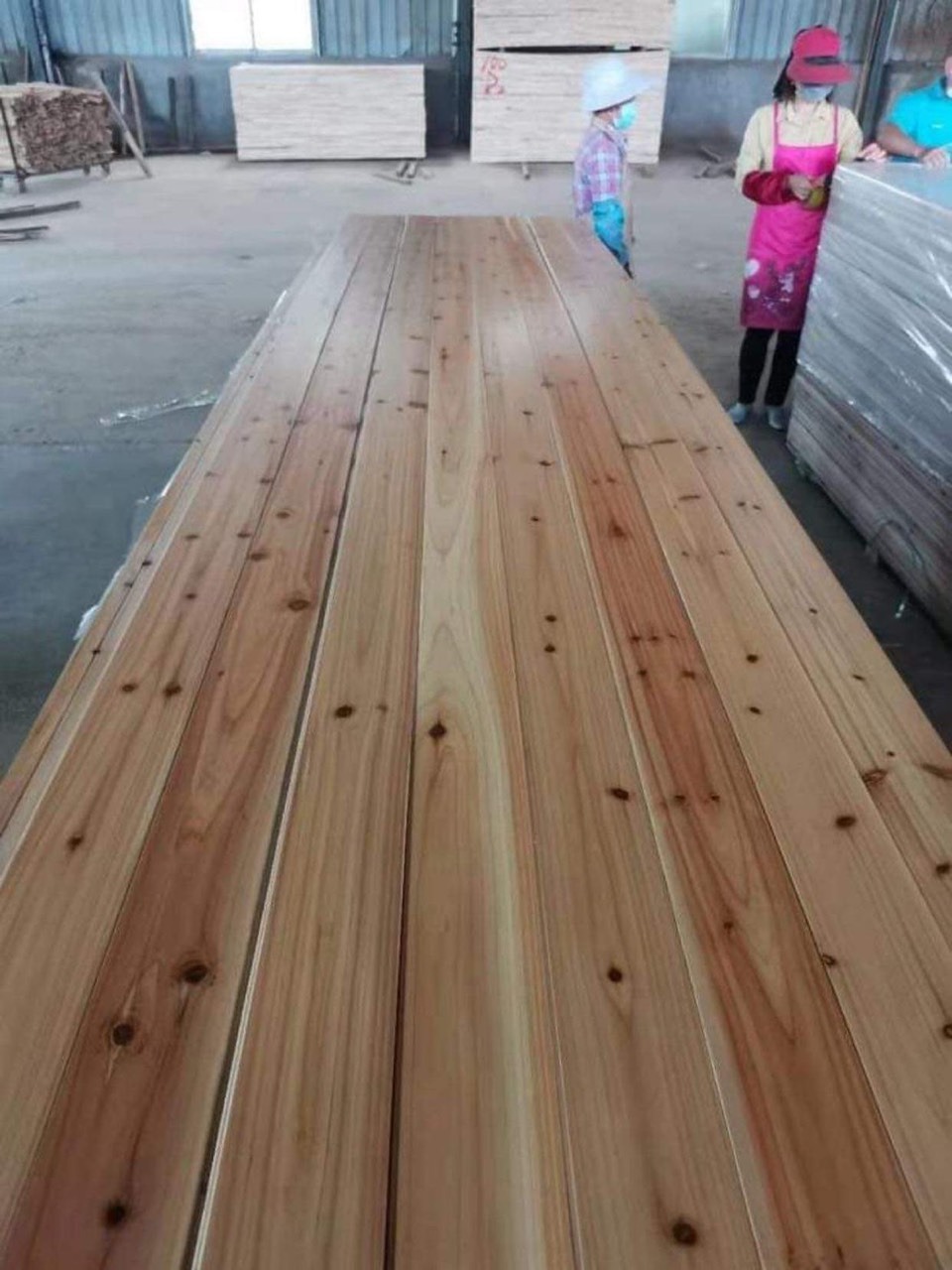 san-xuat-noi-that-va-kien-truc Tấm gỗ dài trong xưởng sản xuất, công nhân đứng cạnh, ánh sáng tự nhiên chiếu vào từ cửa sổ.