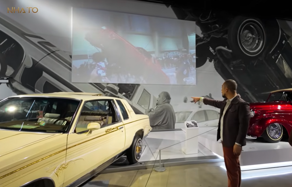 screenshot-2025-07-08-154502 Người đàn ông chỉ vào tấm hình chiếu ô tô cổ điển trong bảo tàng xe hơi.