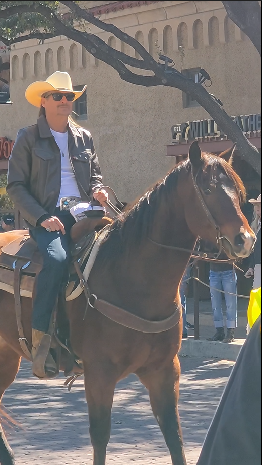 screenshot-2025-07-17-163415 Người đàn ông đội mũ cao bồi cưỡi ngựa trên phố, mặc áo khoác da và kính mát, khung cảnh thành phố phía sau.