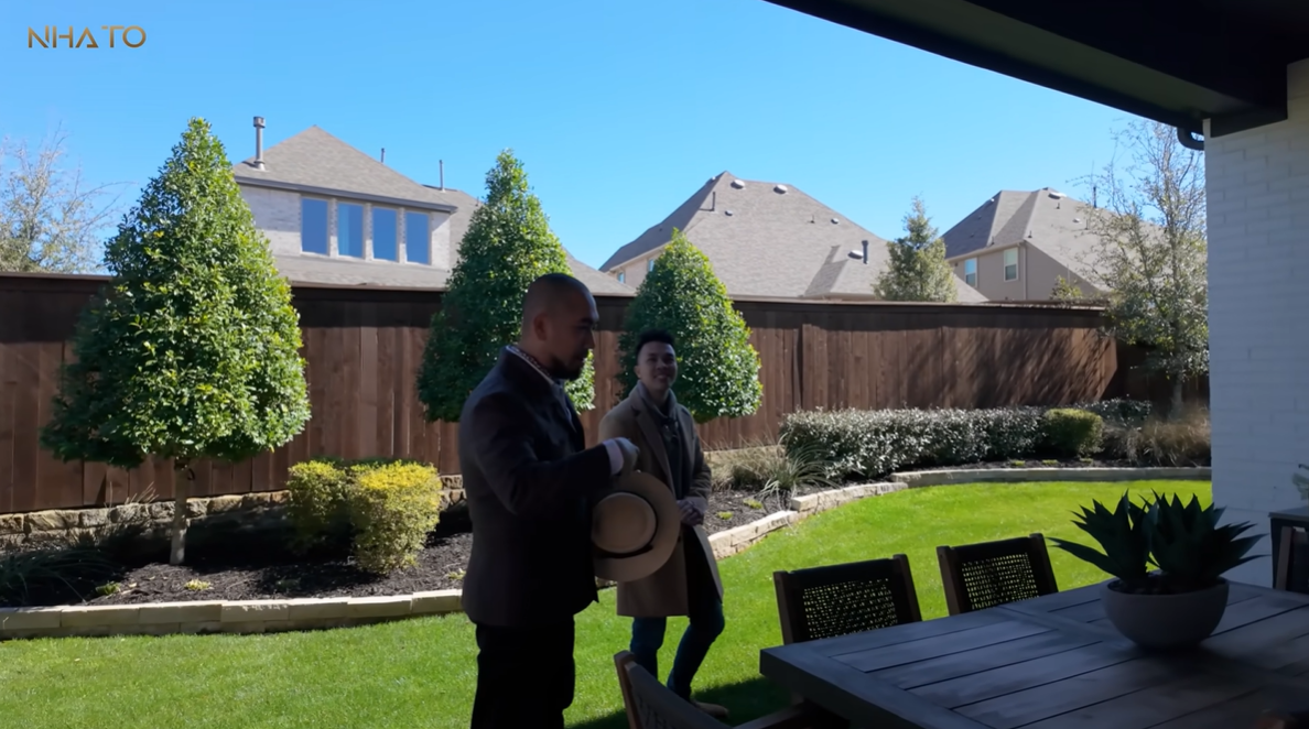 Người đàn ông trò chuyện trong sân vườn nhà, có bàn ghế gỗ và cây xanh bao quanh, trời quang đãng.