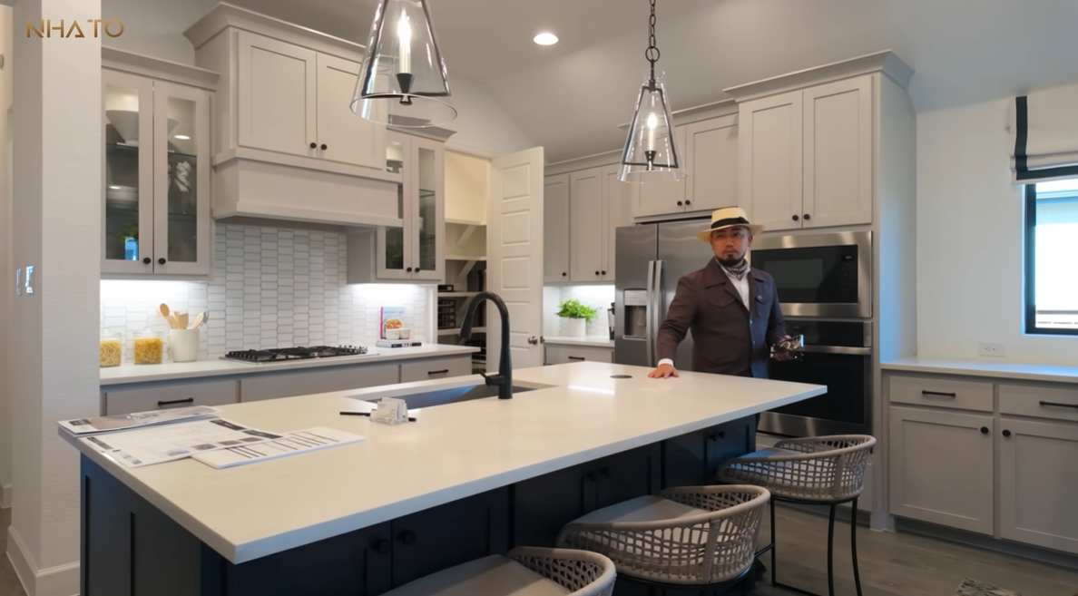 screenshot-2025-07-21-194905-1 Người đàn ông đứng trong nhà bếp hiện đại với tủ trắng và đèn treo trang trí.